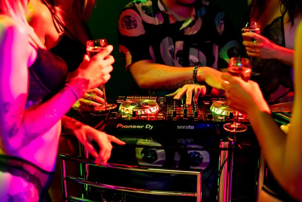 DJ aux platines entouré de convives verre à la main sous des lumières roses au L’Ôlà
